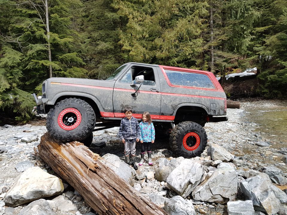 Lifted Ford Bronco with red trim and wheels parked on rocks near a stream, two kids standing next to it