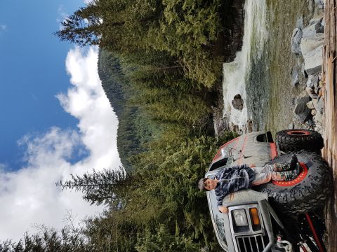 1984 Ford Bronco - A person rests on the hood of a modified off-road vehicle beside a waterfall in a wooded area.