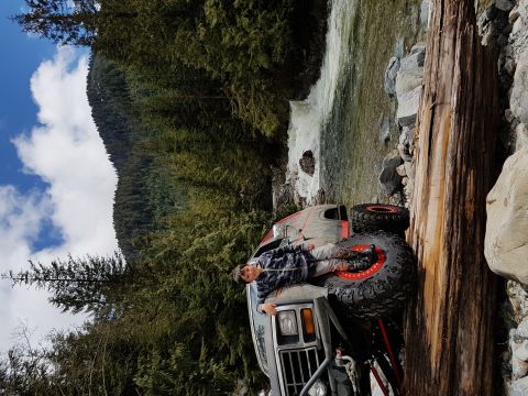 1984 Ford Bronco - A person rests on the hood of a modified off-road vehicle near a waterfall in a wooded area.
