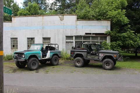 1972 Ford Bronco - Two Ford Broncos, one green and one dark gray, are parked in front of a weathered building with broken windows.