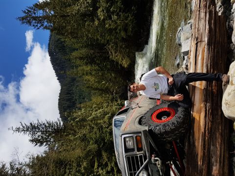 1984 Ford Bronco - A man poses next to a lifted 1984 Ford Bronco parked on a large log next to a river with a waterfall.