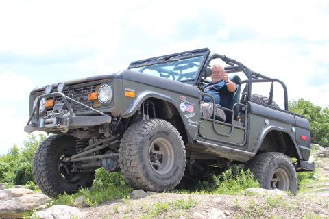 1972 Ford Bronco - A man gives a thumbs-up from the driver's seat of a lifted, olive-drab Ford Bronco with a roll cage and off-road tires. The vehicle is park…