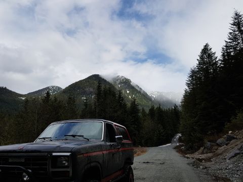 1984 Ford Bronco - A dark colored SUV with red trim is parked on a gravel road in a mountainous, forested area. The vehicle is facing the camera with the fron…
