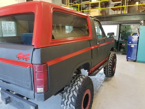 1984 Ford Bronco - The rear passenger side of a dark gray 1980s Ford Bronco with a red roof and red racing stripes is shown in a garage or workshop. It featur…