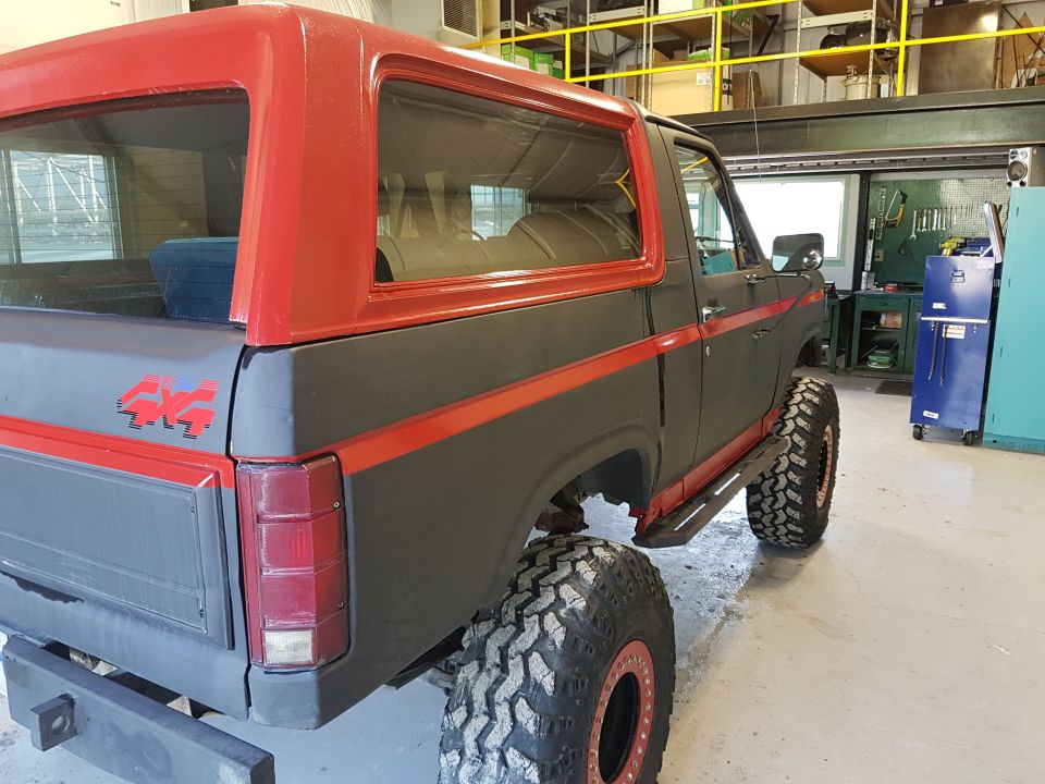 1980s Ford Bronco, dark gray with red roof and stripes, large off-road tires, 4x4 decal, in a workshop.