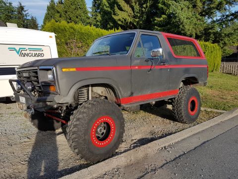 1984 Ford Bronco - A lifted, dark gray 1980s Ford Bronco with red accents is parked on a gravel shoulder next to a paved road, with a white RV in the backgrou…