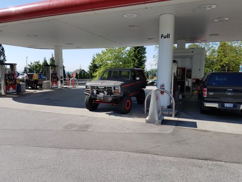 1984 Ford Bronco - A lifted, dark gray 1980s Ford Bronco with red accents is parked at a gas station under a red and white canopy. A black Ford F-150 pickup t…