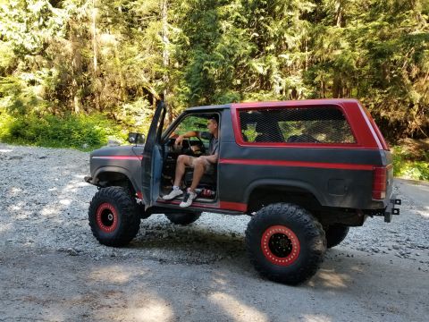 1984 Ford Bronco - A man sits in the driver's seat of a modified black and red Ford Bronco with large off-road tires parked on a gravel road in a forest.