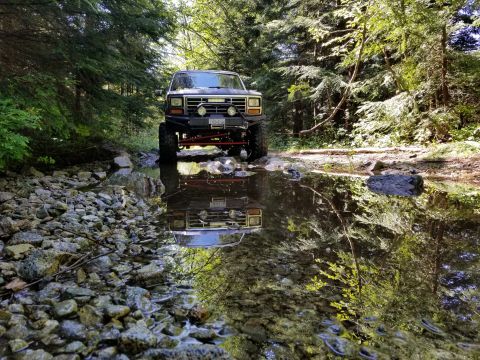 1984 Ford Bronco - A lifted, dark-colored Ford Bronco is parked in a shallow, rocky stream bed surrounded by trees. The front of the vehicle and its reflectio…