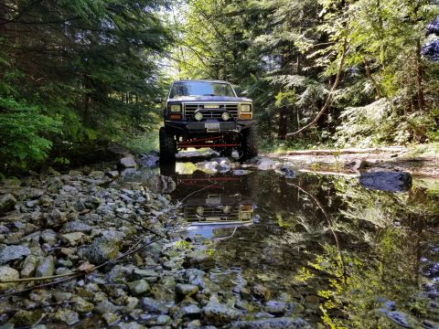 1984 Ford Bronco - A lifted, dark-colored 1980s Ford Bronco is parked in a shallow, rocky stream bed surrounded by trees. The front of the vehicle is reflecte…