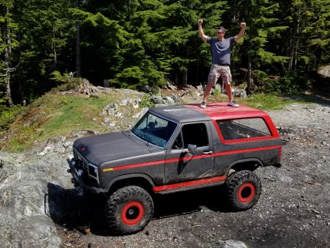 1984 Ford Bronco - A man stands triumphantly on the roof of a lifted, dark gray Ford Bronco with a red top, off-roading on a rocky, wooded trail.