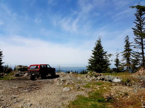 1984 Ford Bronco - A red 1984 Ford Bronco is parked on a rocky, mountainous terrain with pine trees and a distant city visible under a blue sky with wispy clo…