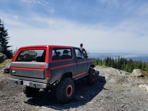 1984 Ford Bronco - A lifted, dark gray and red 1980s Ford Bronco is parked on a rocky, wooded hilltop overlooking a distant city and bay. A person sits on the…