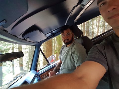 1984 Ford Bronco - Two men are inside the cab of a blue Ford Bronco, looking out at a forest. The interior shows a dark blue headliner and a tan dashboard.