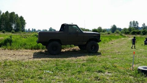1984 Ford Bronco - A dark-colored, lifted 1980s Ford Bronco is parked in a grassy field with trees in the background. A young person walks away from the vehic…