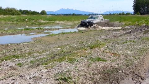 1984 Ford Bronco - A pickup truck navigates a muddy off-road course, splashing through water and dirt.
