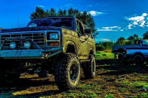 1984 Ford Bronco - A heavily muddied SUV sits in a field under a bright blue sky with clouds.