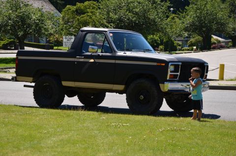 1984 Ford Bronco - A black Ford Bronco with a tan stripe is parked on a grassy area next to a road. A young boy stands near the front bumper.