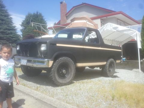 1984 Ford Bronco - A young boy stands next to a dark-colored Ford Bronco with off-road tires in a residential driveway.
