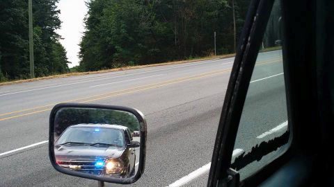 1984 Ford Bronco - A police SUV with its emergency lights activated is visible in the side mirror of another vehicle on a two-lane road.