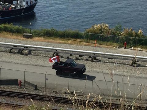 1984 Ford Bronco - A black pickup truck drives along a road next to train tracks, with a Canadian flag attached to its rear.