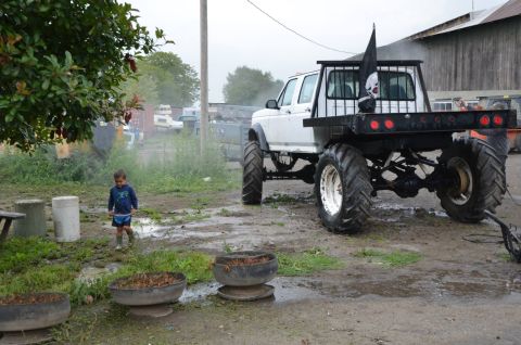 1984 Ford Bronco - A modified pickup truck with large tires and a pirate flag is parked in a muddy yard next to a child.