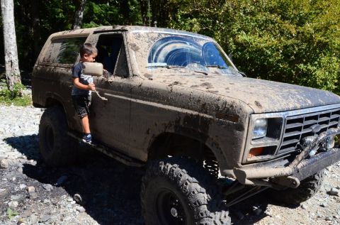 1984 Ford Bronco - A young boy stands by a heavily mud-caked SUV on a rocky, wooded trail.