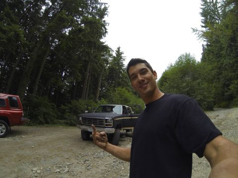 1984 Ford Bronco - A man stands in front of a pickup truck and a red SUV in a wooded, off-road setting.