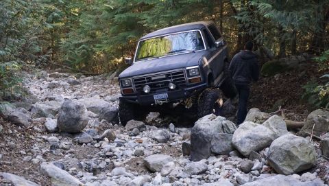 1984 Ford Bronco - A dark blue 1984 Ford Bronco tackles a challenging rocky terrain in a wooded area.