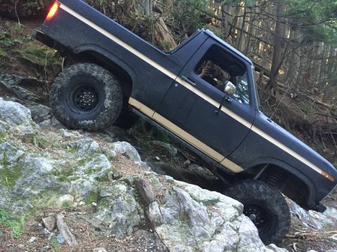 1984 Ford Bronco - This SUV is navigating a challenging off-road trail with large rocks and trees.