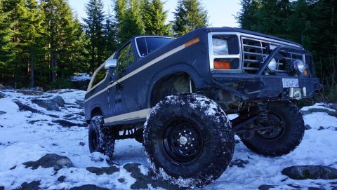 1984 Ford Bronco - A modified dark blue SUV with large tires navigates a snowy, rocky path through a forest.