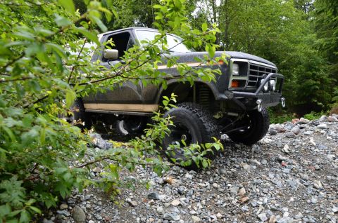 1984 Ford Bronco - A lifted Ford pickup truck navigates a rough, wooded terrain with dense foliage obscuring parts of the vehicle.