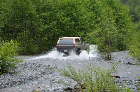 1984 Ford Bronco - A 1984 Ford Bronco navigates a rocky riverbed, kicking up water as it drives through the natural obstacle.