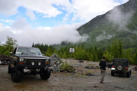 1984 Ford Bronco - Two off-road vehicles are parked in a mountainous, wooded area with cloudy skies.