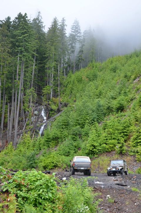 1984 Ford Bronco - Two SUVs navigate a forest road, with a waterfall cascading down a rocky slope.