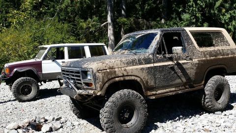 1984 Ford Bronco - Two off-road vehicles, caked in mud, sit parked on a rocky trail surrounded by dense forest.