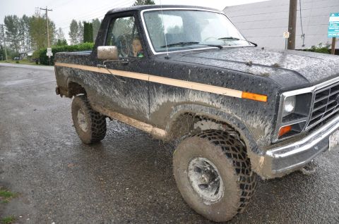 1984 Ford Bronco - A child looks out the window of a muddy black Ford Bronco driving on a wet road.