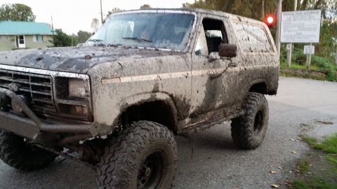 1984 Ford Bronco - This heavily mud-caked SUV, likely from the 1980s, is shown from a three-quarter front perspective.