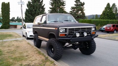 1984 Ford Bronco - A modified black Ford Bronco is parked on the side of the road next to a white minivan.