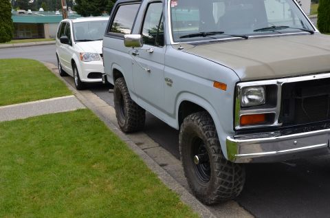 1984 Ford Bronco - A light blue Ford Bronco is parked on the street next to a white minivan.