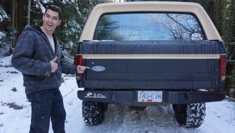 1984 Ford Bronco - A man points to the Ford badge on the rear of a 1984 Ford Bronco, parked in a snowy, wooded area.