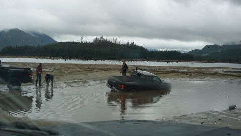 1984 Ford Bronco - A black pickup truck is stuck in shallow water with two people attempting to help.