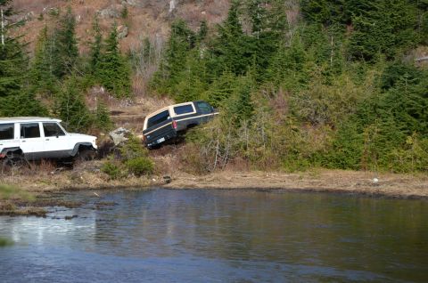 1984 Ford Bronco - Two SUVs navigate a challenging off-road terrain near a body of water, with one vehicle partially on a steep, wooded embankment.