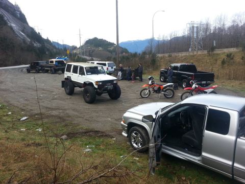 1984 Ford Bronco - Several vehicles, including a white off-road SUV and motorcycles, are parked in a dirt lot near a mountain.