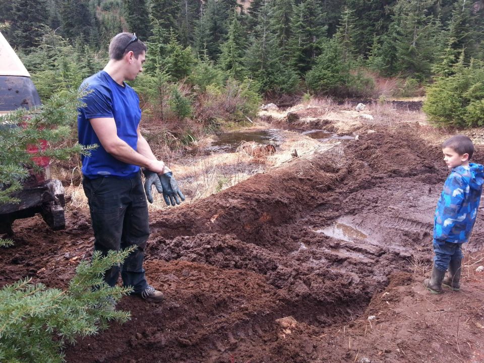 Man in blue shirt and gloves stands near a muddy ditch while a child in a blue jacket watches
