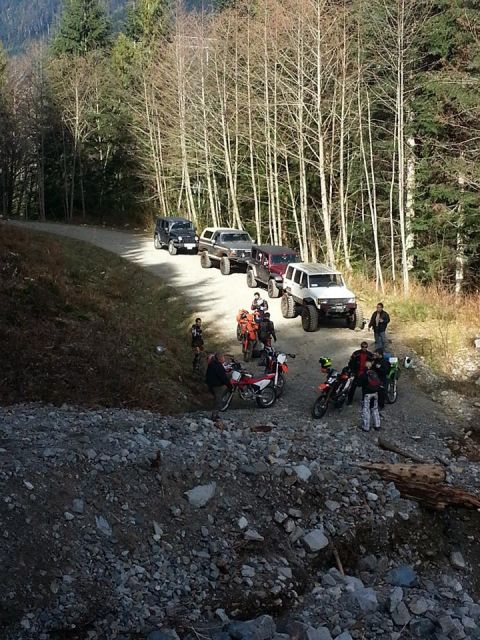 1984 Ford Bronco - A group of off-road vehicles and motorcycles are stopped on a dirt road surrounded by trees.