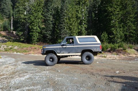 1984 Ford Bronco - A lifted Ford Bronco drives on a gravel road through a wooded area.
