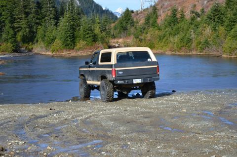 1984 Ford Bronco - This 1984 Ford Bronco, modified with a lifted suspension, is positioned near the edge of a river with a forested mountain backdrop.