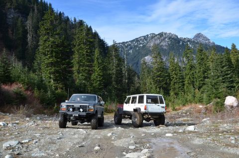 1984 Ford Bronco - Two off-road SUVs, a Ford Bronco and a Jeep Cherokee, are parked on a dirt road in a mountainous, forested area.
