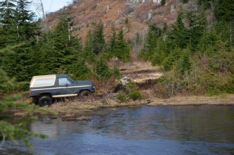 1984 Ford Bronco - A classic Ford Bronco sits off-road near a lake, surrounded by evergreen trees and a rocky hillside.